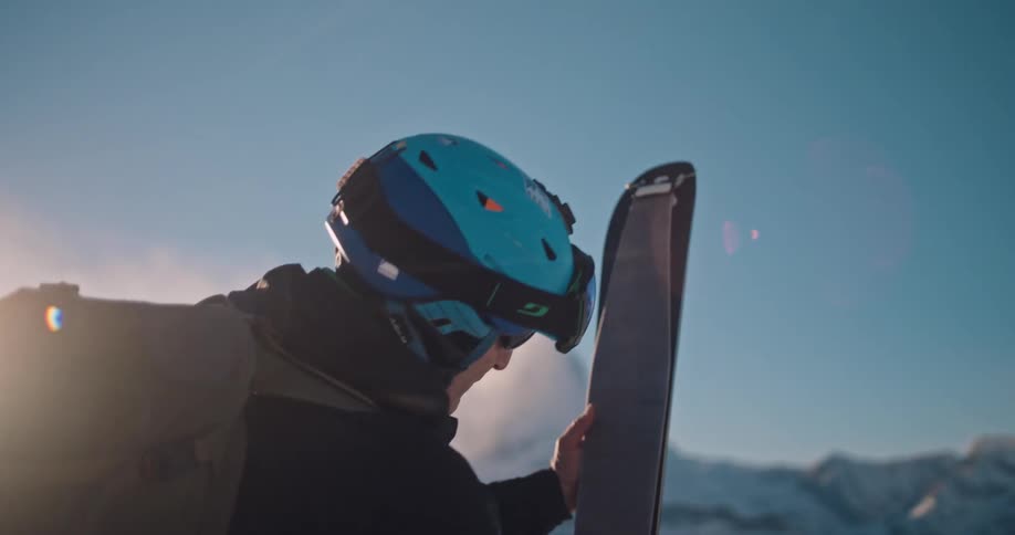 A backcountry skier in full gear removes the climbing skins from his skis on a sunlit mountain peak in slow motion