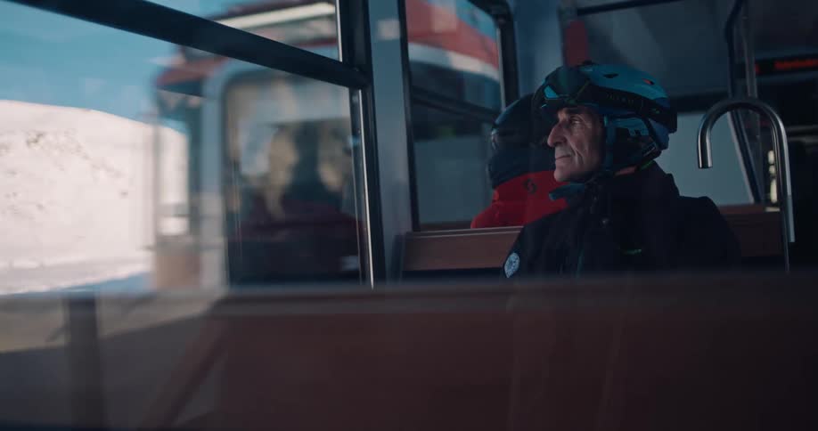 Pensive older skier in a helmet and goggles looks out the window of a mountain train at the bright, snowy landscape passing by