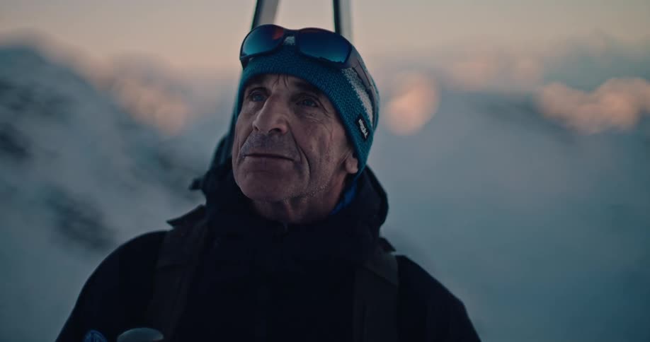 A contemplative senior male skier with skis on his back admires the vast, snowy mountain landscape at sunrise