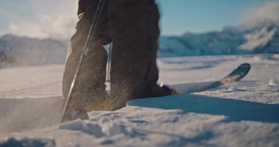 A skier clips into their bindings in slow motion, kicking up fresh powder with sunny, snow-covered mountains in the background