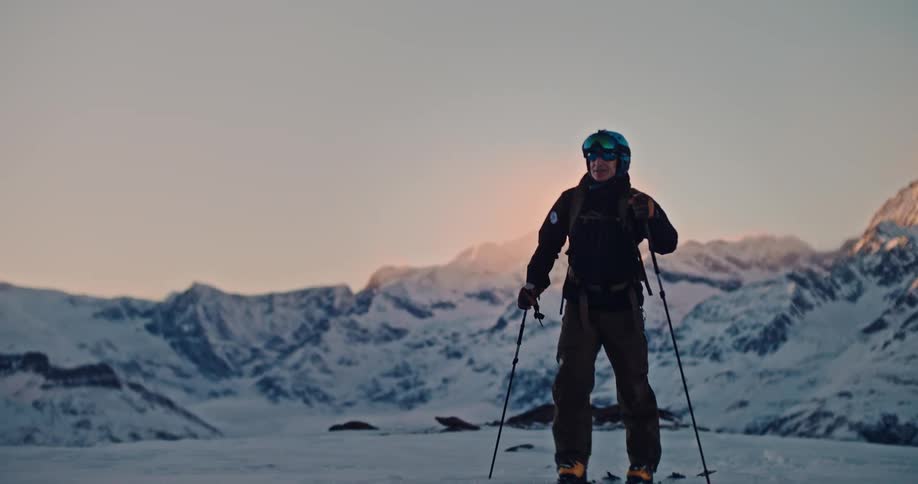 A skier walks towards the camera in slow motion across a snowy landscape with a majestic mountain range illuminated by the sunrise in the background
