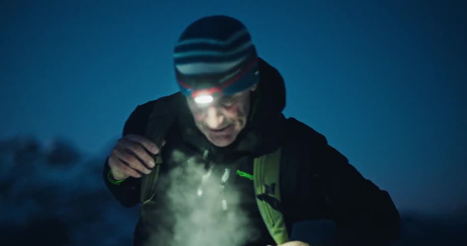 A slow-motion rack focus reveals an elderly hiker with a headlamp putting on his backpack in the cold night