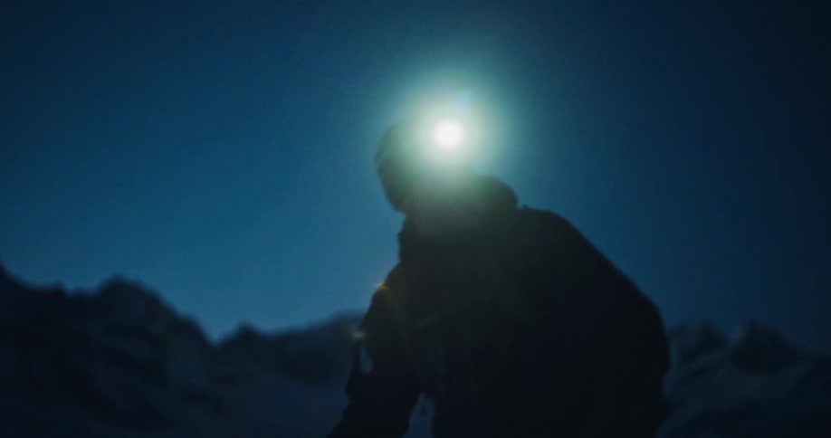 A silhouetted hiker with a bright headlamp makes his way up a snowy mountain in the predawn darkness