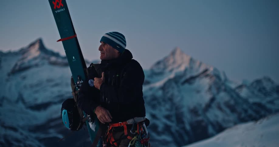 A backcountry skier pauses to admire the alpine view at dusk before taking off his backpack and placing it in the snow