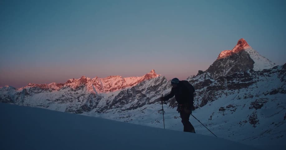 A lone backcountry skier ascends a snowy slope at sunrise, with the iconic Matterhorn illuminated by a beautiful alpenglow