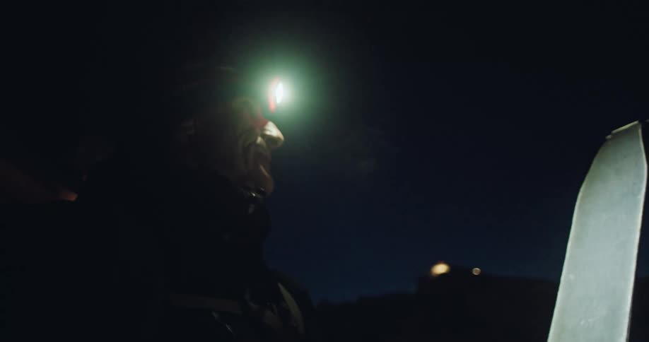 A point-of-view shot from a ski shows a backcountry skier applying climbing skins in the dark, illuminated by his headlamp