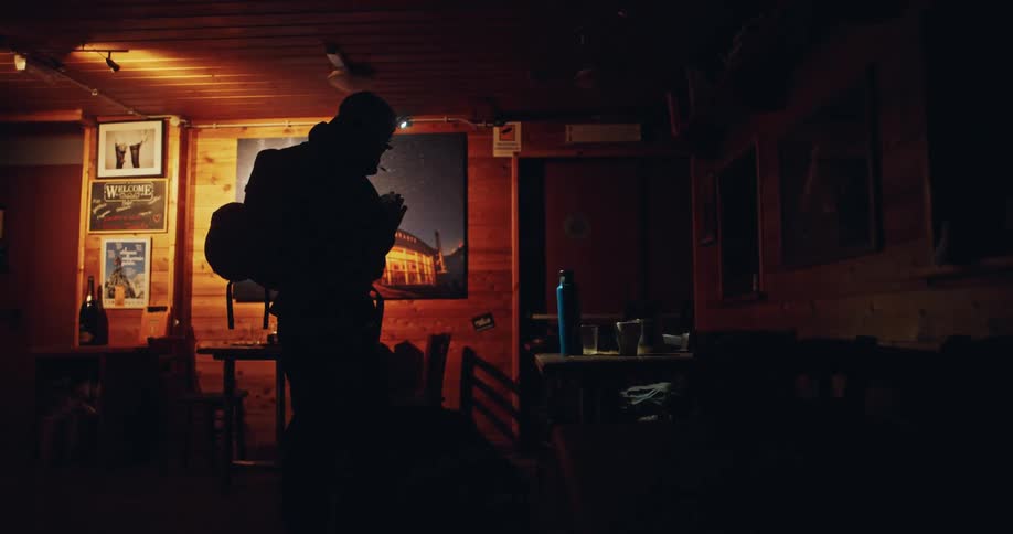 A hiker wearing a headlamp packs his backpack inside a dark wooden cabin before an early morning adventure