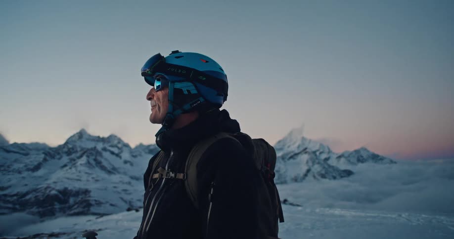 An older skier in full gear takes a moment to admire the epic sunrise view from a snowy mountain peak above the clouds