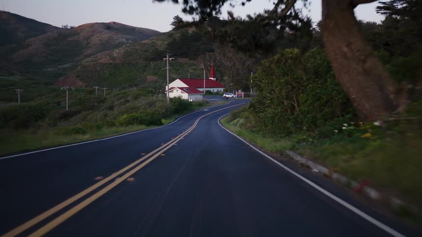 A slow, serene point-of-view drive down a winding country road through rolling green hills towards a small building at dusk