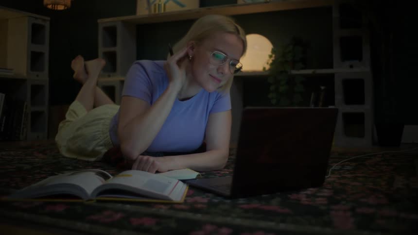 A smiling female student lies on the floor of her cozy room at night, studying with her laptop and textbook