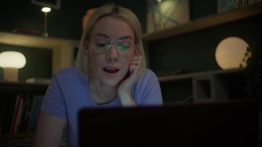 A focused young woman wearing glasses has a surprised reaction while looking at her laptop screen at night