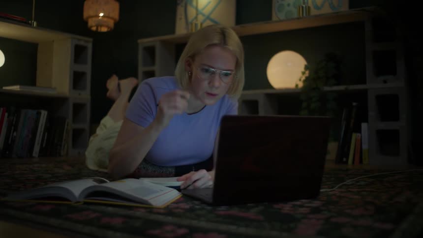 A focused female student with glasses studies late into the night, taking notes from her laptop while comfortably lying on the floor