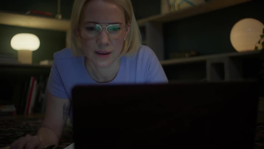 A focused blonde woman with glasses lies on the floor while studying on her laptop late at night in her living room
