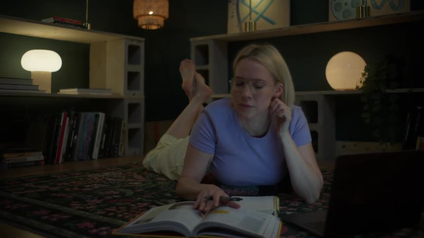 A focused young woman with glasses studies late into the night, lying on the floor with her laptop and books in a cozy, dimly lit room