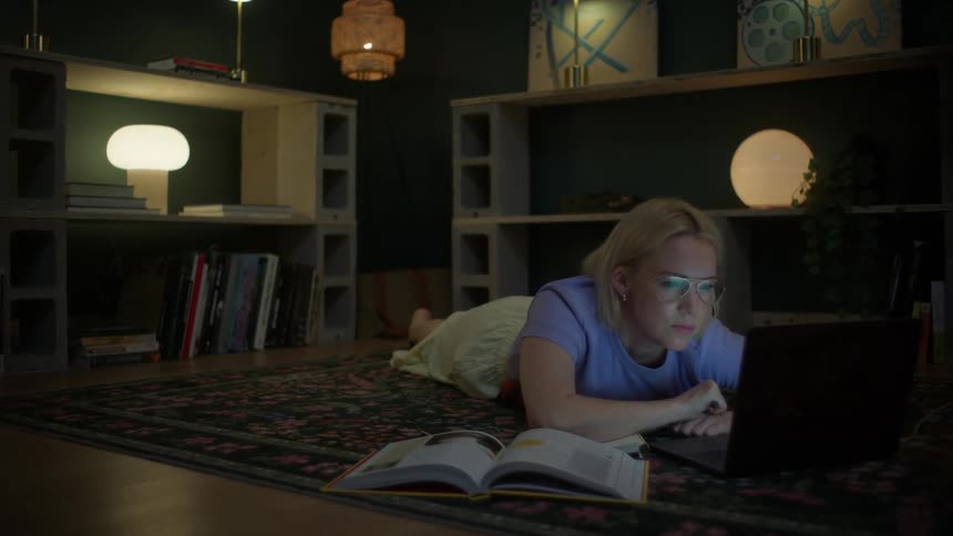 In a dimly lit and cozy room, a tired young woman with glasses dozes off while studying late at night on her laptop on the floor
