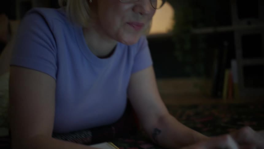 A focused blonde woman wearing glasses uses her laptop at night while comfortably lying on the floor