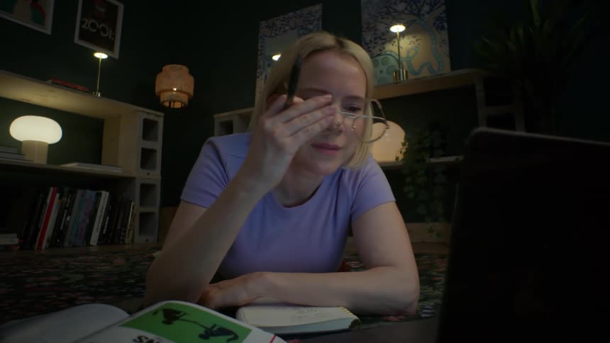 A tired female student rubs her eyes and puts on her glasses while studying late at night on her laptop in a dimly lit room