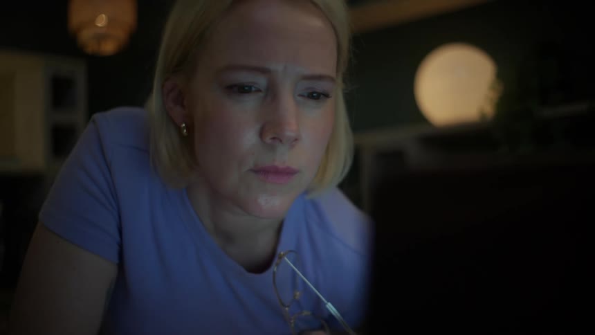 A concerned blonde woman works late into the night, her face illuminated by the laptop screen as she scrolls with a pensive expression