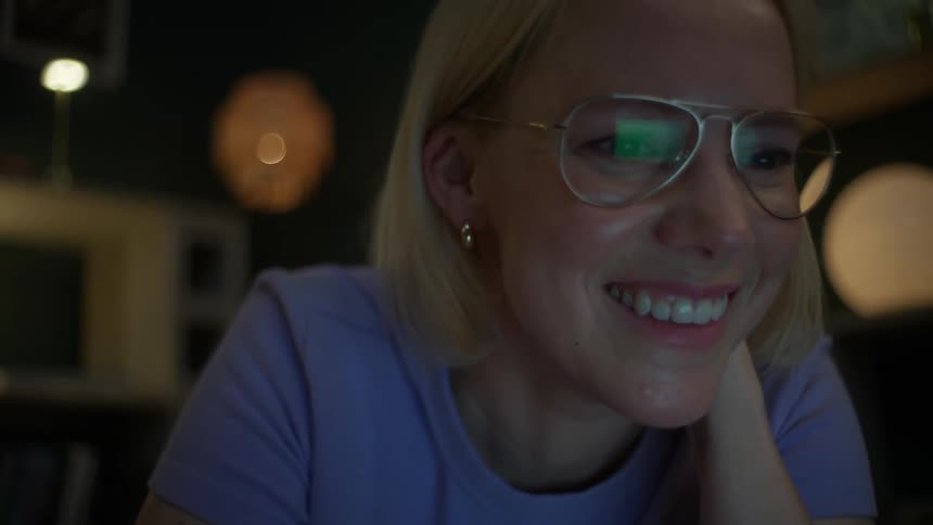 A woman with glasses studying on the floor at night smiles brightly as she looks at something on her laptop screen
