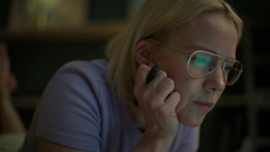 A woman with glasses thoughtfully works on her laptop late at night in a dimly lit room