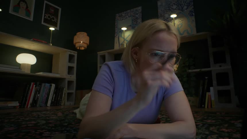 A dedicated student wearing glasses studies late into the night, using her laptop and books while lying on the floor of her cozy apartment