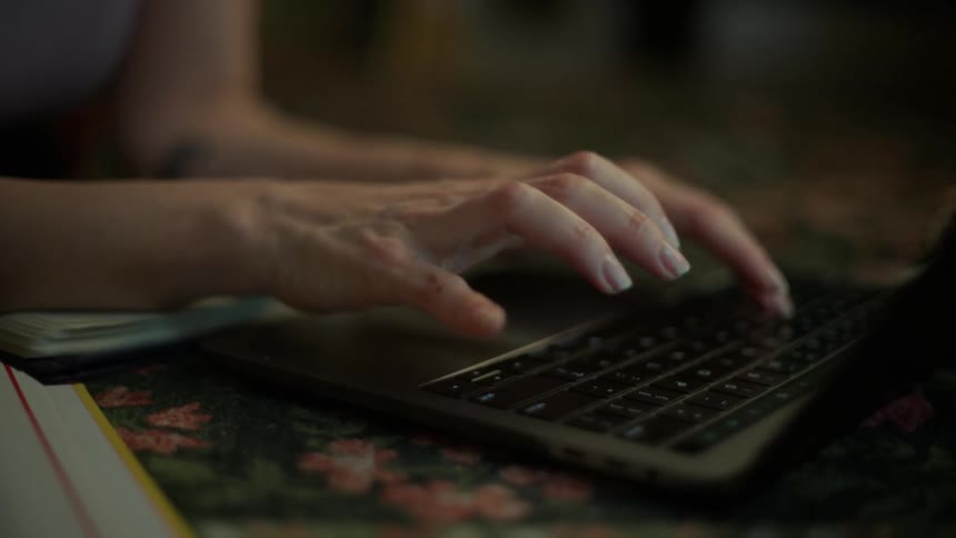 Close-up of a woman's hands typing on a laptop keyboard in a dimly lit, moody room