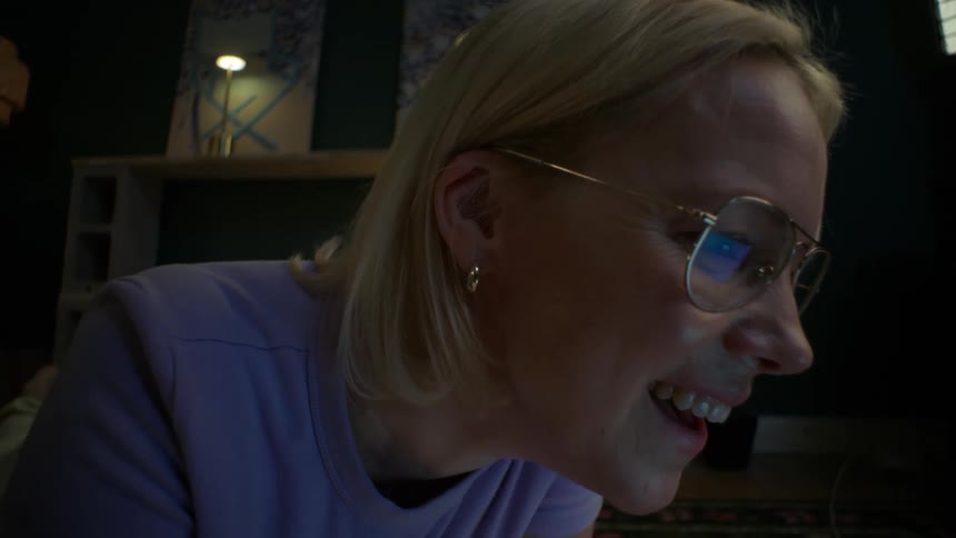 A smiling college student with glasses researches on her laptop at night, lying on the floor surrounded by books in her cozy apartment