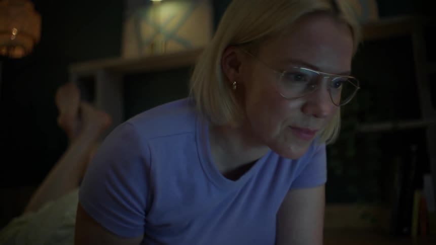 A blonde woman in glasses comfortably lies on the floor at night, writing in a notebook while studying with a laptop and books