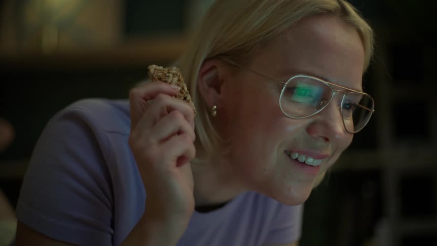 A woman in glasses smiles as she eats a granola bar while looking at her laptop in a dimly lit room