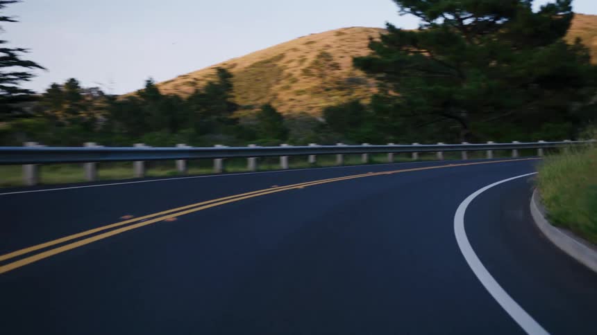 A smooth point-of-view shot driving along a winding country road through lush green hills and forests at sunset