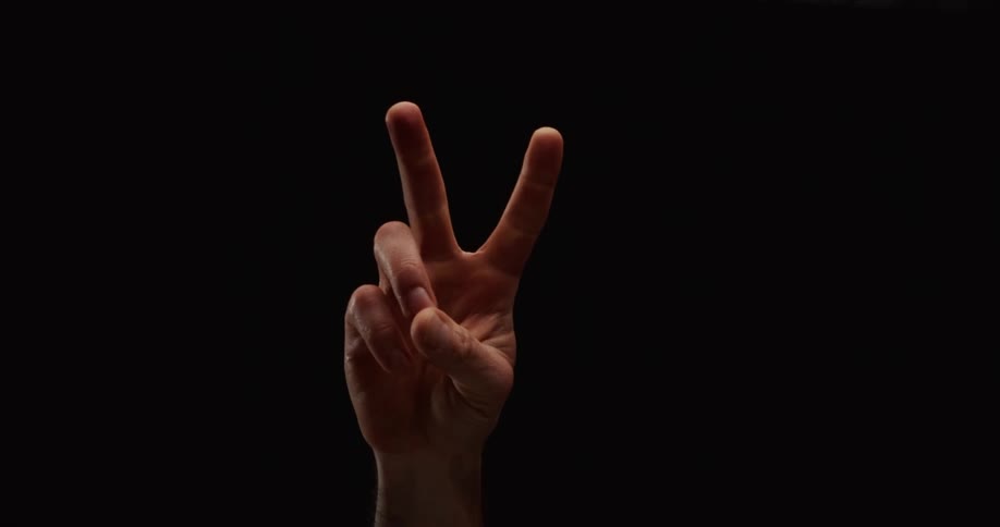 A male hand rises from the bottom of the frame and makes a peace sign against a black background