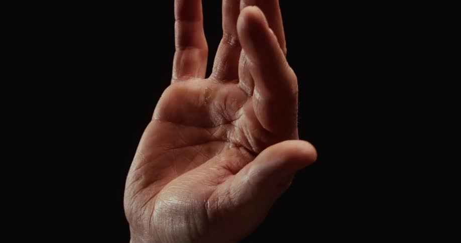 A dramatic closeup of a male hand slowly unfurling its fingers against a black background
