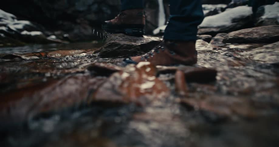 A slow-motion close-up of a hiker's boots splashing as they carefully cross a rocky stream in a winter landscape