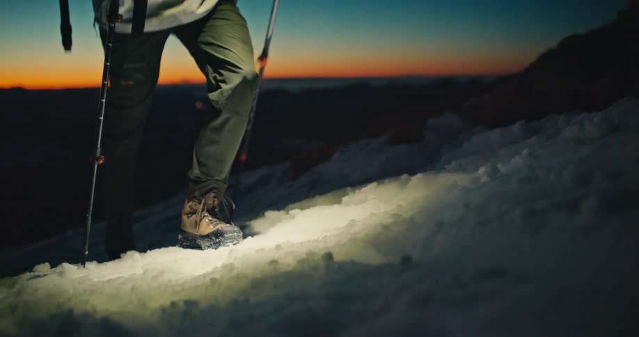 A lone mountaineer ascends a snowy peak in slow motion at sunrise, their headlamp illuminating the path forward