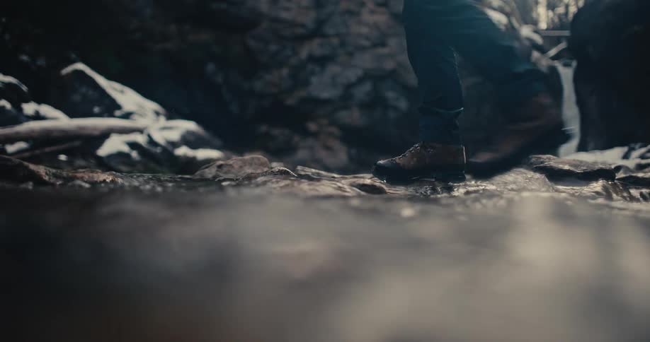 Slow motion, low angle view of a person in hiking boots carefully stepping through a shallow, rocky stream with a waterfall in the distance
