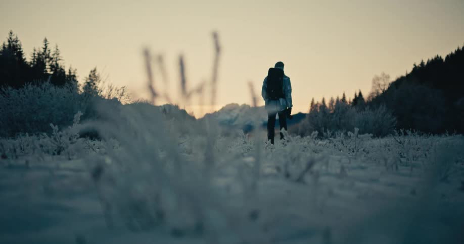 A solitary photographer with a backpack walks through a frost-covered snowy meadow towards the mountains at sunrise in slow motion