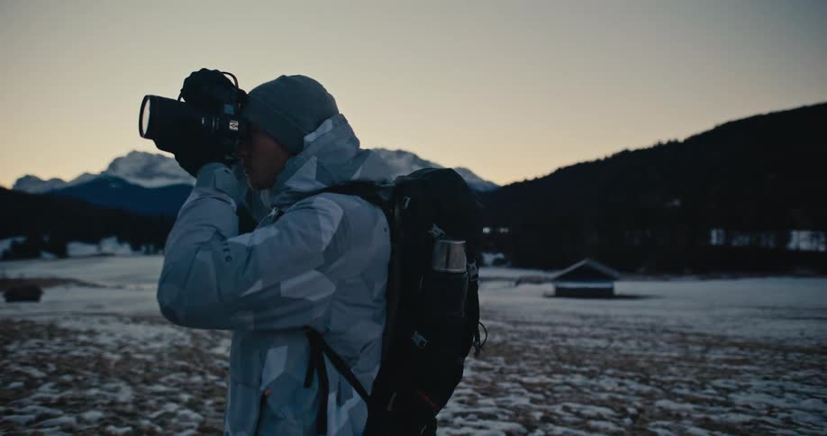 A male photographer in winter gear frames a shot of the snowy mountain landscape at dusk