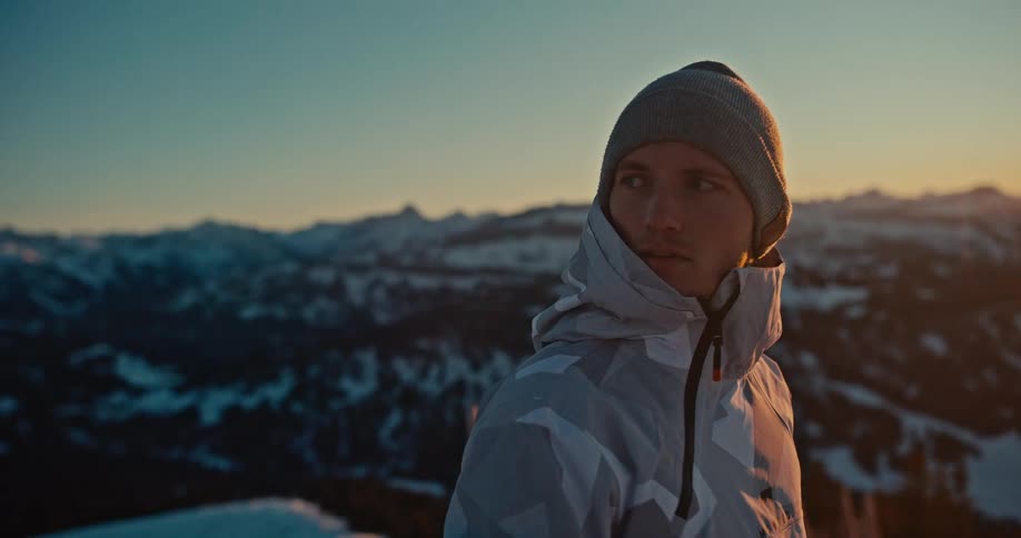 A pensive man in winter gear watches the golden sunset from a snowy mountaintop, overlooking a vast alpine range