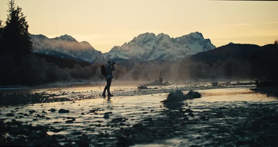 A solo traveler and photographer walks along a misty river to capture the stunning mountain landscape at sunrise