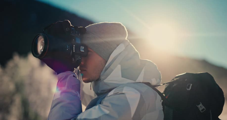 A professional photographer dressed in winter gear raises his camera to capture a shot in the bright, cold sunlight