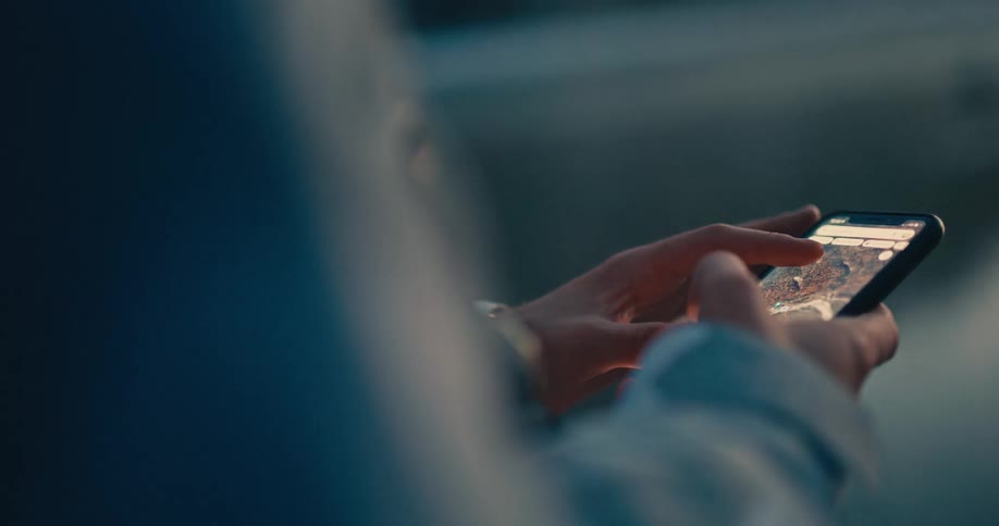 Close-up over-the-shoulder shot of hands scrolling and zooming on a satellite map on a smartphone at dusk