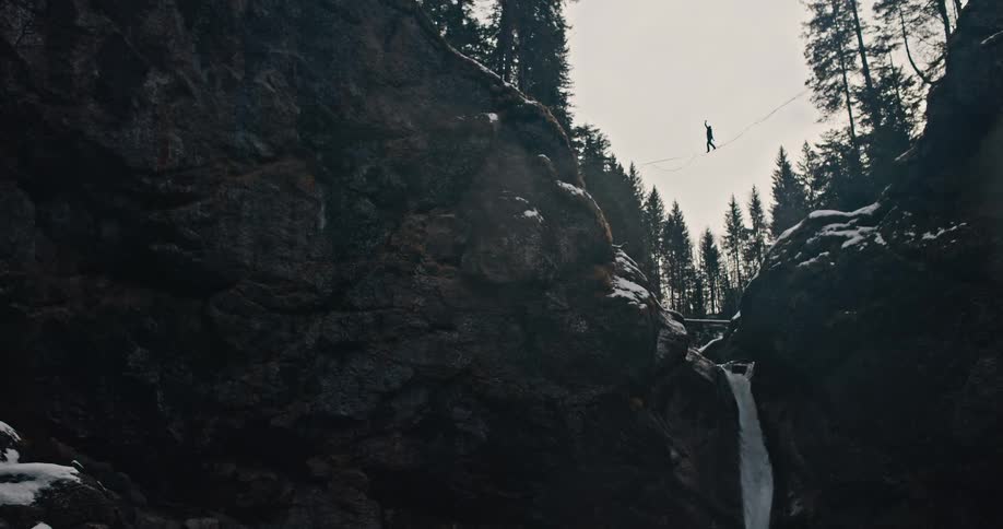 A brave person slacklines across a highline suspended over a waterfall in a dramatic and snowy mountain gorge