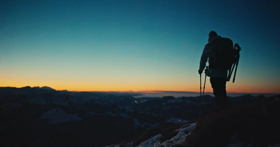 A hiker with a headlamp and poles reaches a snowy mountain summit at twilight, stopping to admire the vast landscape
