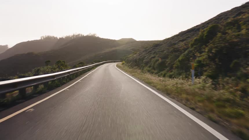 A scenic point-of-view drive along a winding mountain road surrounded by green hills under a bright, hazy sky