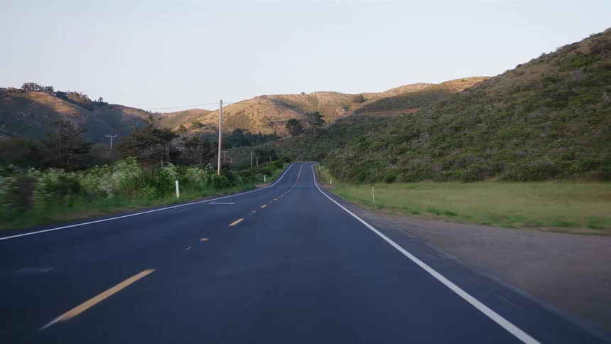 A serene point-of-view drive along an empty rural highway through rolling green hills at dusk