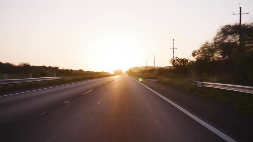 A point-of-view shot driving down an empty rural highway into the warm, glowing sunrise