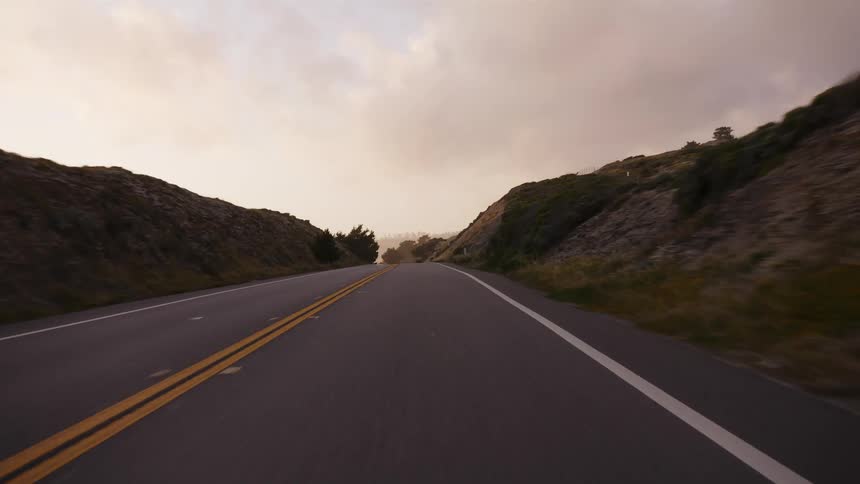 A smooth point-of-view shot driving down a scenic highway flanked by rolling hills under a warm, cloudy sky