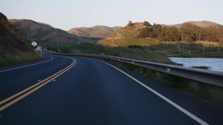 Driving POV along a winding scenic road next to a lake with rolling green hills in the background during golden hour