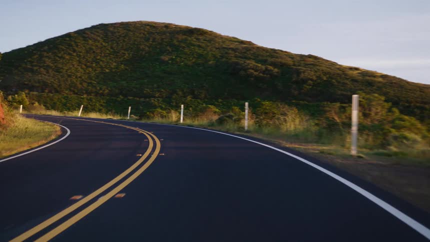A slow, smooth point-of-view drive along a winding country road through rolling green hills during golden hour