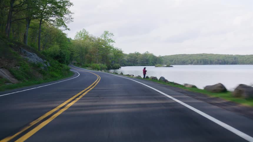 Driving POV on a winding lakeside road in the forest, passing a person fishing and a crashed car